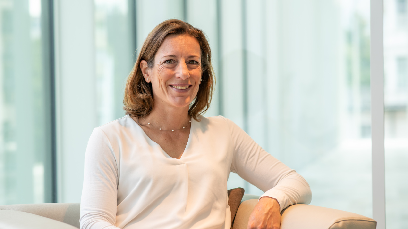 A portrait of Nicola Spirig. She is wearing a white long-sleeved shirt and is sitting in a beige armchair.