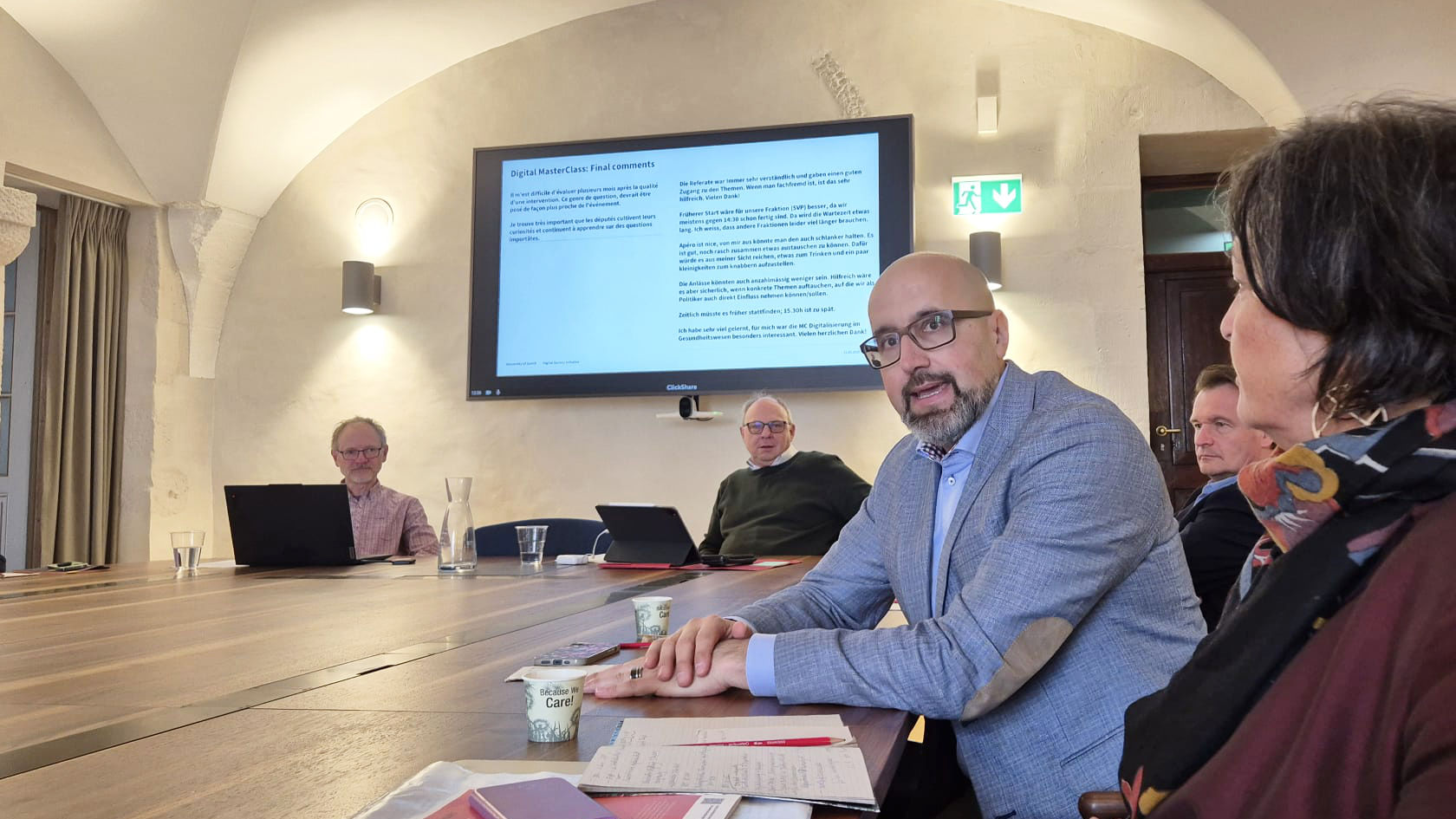 Fünf Personen Sitzen um einen Tisch, im Hintergrund ist ein BIldschirm mit einer Präsentation zu sehen.