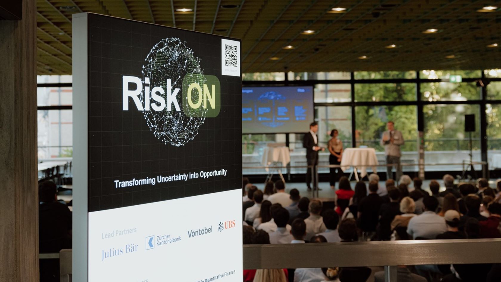 Schild mit der Aufschrift "RiskON" vor einem Saal mit zahlreichen Leuten