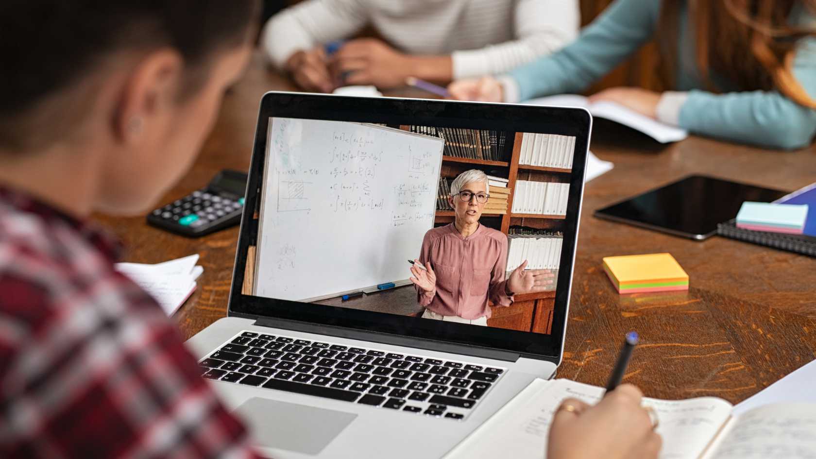 View over the shoulder of a student looking at a laptop on the table in front of her. The laptop is displaying a video of a course.