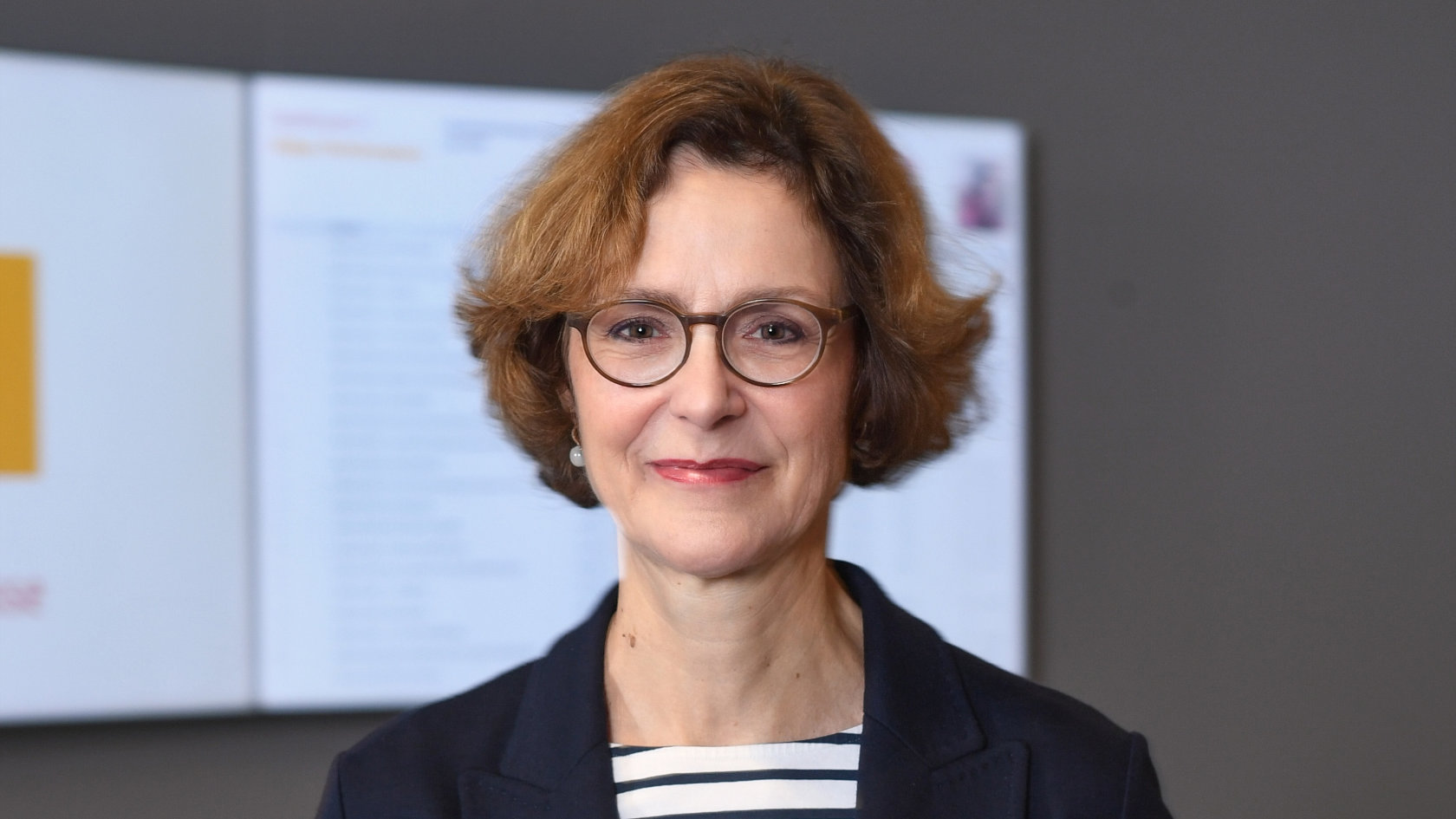 Monika Rühl mit kurzen braunen Haaren, Brille und dunkelblauem Blazer vor unscharfem Bildschirm im Hintergrund